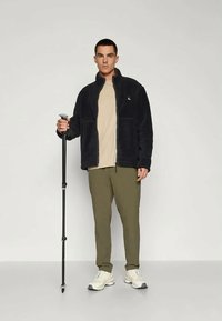 Crna flis jakna, bež majica, maslinasto zelene hlače i bijele tenisice. Drži crni planinarski štap, stoji na neutralnoj pozadini.