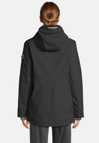 Persoon met opgestoken haar die een zwarte hoodie-jack draagt, van achter gezien, met grijze broek en zichtbaar gestreepte mouwen.