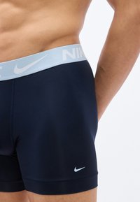 Άνδρας που φοράει σκούρα μπλε boxer briefs της Nike με ανοιχτό μπλε ζώνη μέσης με το λογότυπο της Nike, εμφανιζόμενος από τη μέση μέχρι το μέσο μηρού σε λευκό φόντο.