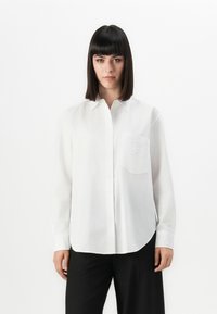Woman with straight dark hair wearing a white button-up shirt with a chest pocket and black wide-leg trousers, standing against a white background.
