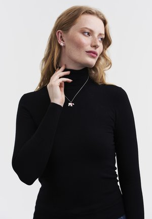 Jauna raudonplaukė moteris juodu golfu, liesianti savo kaklą, demonstruojanti baltos spalvos meškiuko pakabuko vėrinį ir derančius meškiuko formos auskarus.