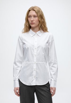 Femme aux longs cheveux blonds ondulés, portant une chemise blanche à manches longues boutonnée et un pantalon foncé, debout devant un fond clair uni.
