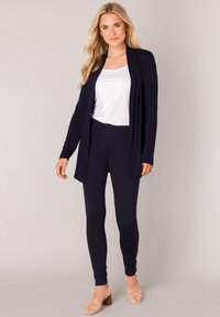 Marineblauwe open-front cardigan over een witte top en bijpassende legging, met een zachte textuur en een casual pasvorm. Sleehakken met bandjes maken de look compleet.