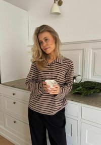 Prugasta smeđa i bež košulja dugih rukava s kragnom, uparena s crnim hlačama; drži bijelu zdjelu nasuprot svijetloj kuhinjskoj pozadini.