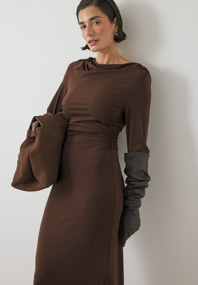 Abito lungo marrone a maniche lunghe con vita arricciata, con guanti in pelle nera e una borsa marrone a texture morbida con chiusura a zip.
