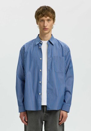Jeune homme portant une chemise rayée bleu et blanc avec des boutons par-dessus un t-shirt blanc et un pantalon gris, debout devant un fond blanc.