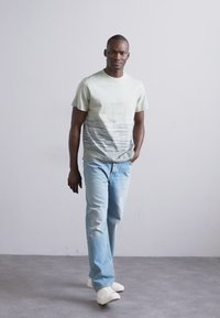Light green t-shirt with a gradient pattern transitioning to gray, paired with light blue jeans and white sneakers, worn by a male model.
