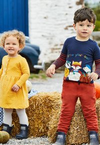 Dvoje djece stoji na slami, jedno u haljinici s žutim točkicama, a drugo u tamnoplavoj majici s grafikom životinja i smešnim hlačama, oboje u crnim čizmama.