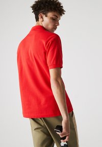 Camisa polo roja de manga corta, de color sólido, material de algodón, con cuello plano, combinada con pantalones beige, sosteniendo un cinturón negro en la mano.