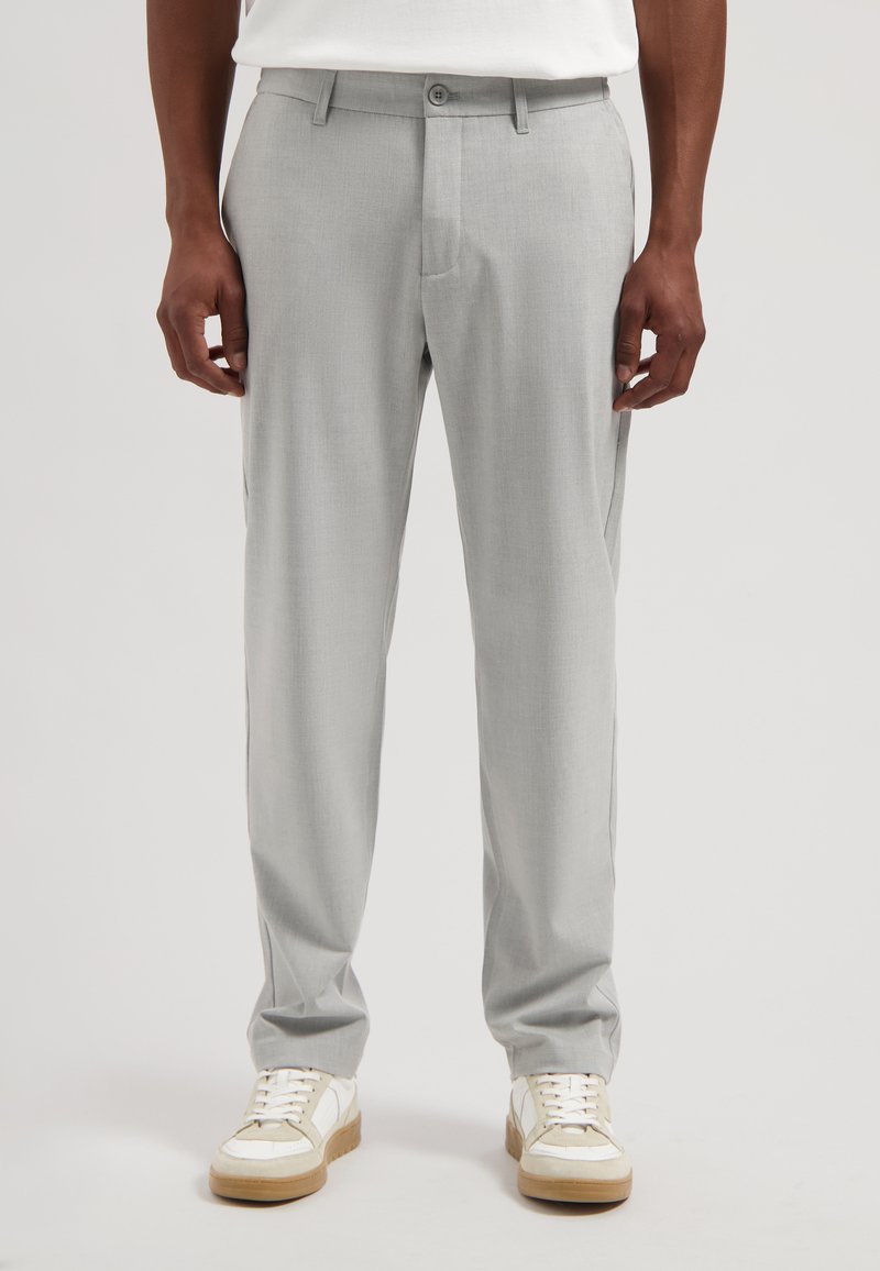 Pantalon gris clair sur mesure en tissu lisse. Dispose d'une fermeture à bouton, de poches latérales et d'une coupe slim. Associé à des baskets blanches.
