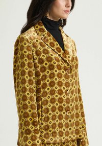 Blazer in raso giallo con motivo geometrico marrone, dotato di colletto a risvolto, bottoni frontali e texture liscia.
