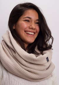 Beige knitted infinity scarf with a textured pattern and a small logo tag, worn draped around the neck, complementing a light sweater.