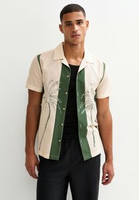 Camicia a maniche corte in tessuto beige e verde con design di palme, strisce verticali verdi e chiusura con bottoni. Maglietta nera sottostante visibile.