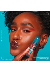 Femme aux cheveux bouclés foncés et maquillage audacieux tenant un gloss teinté hydratant NYX Lip IV près de ses lèvres sur un fond bleu vif.