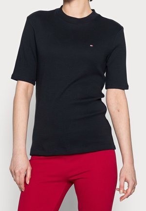 Personne portant un t-shirt noir uni à manches courtes avec un petit logo sur la poitrine et un pantalon rouge vif sur un fond uni.