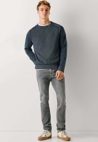 Mladý muž stojaci s rukou v kapse, oblečený v tmavomodrej mikine, svetlošedých džínsoch a béžových teniskách s gumovou podrážkou.