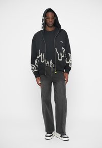 Fekete cipzáras kapucnis pulóver fehér lángmintával, fekete pólóval, szürke farmerrel és fehér, fekete részletekkel ellátott sportcipővel párosítva.