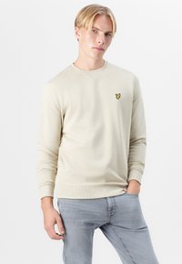 Bežinė crew neck džemperis su įrengtomis rankogaliais ir apatine dalimi, turintis mažą geltoną siuvinėtą logotipą ant krūtinės. Minkšta medžiagos tekstūra.