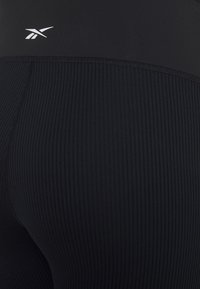 Black ribbed leggings with elastic waistband, featuring a smooth top section and a small white logo on the upper left side.