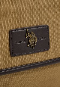 Ruskea kangaskassi, jossa on musta nahkainen merkki. Kullanvärinen tunnus esittää ratsastajahahmoa. Tekstissä lukee "SINCE 1890." Kestävä kanvaskangas.