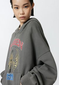 Sudadera gris de gran tamaño con un estampado gráfico que presenta "REINA EN EL CAMINO", acentos dorados y un bolsillo frontal tipo canguro. Tejido suave y texturizado.