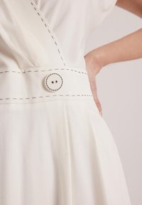 Biała sukienka z widocznymi czarnymi przeszyciami w talii, z okrągłym białym guzikiem. Miękka, lekka tkanina z delikatnym układaniem.