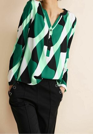 Persoon draagt een groen-wit-zwart blouse met een abstract patroon en lange mouwen, gecombineerd met zwarte broek met knoopdetails bij de zakken.