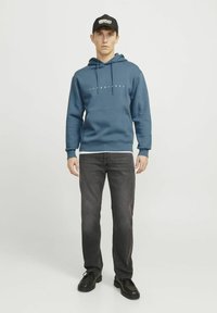 Sininen huppari, jossa on narut ja etutasku, jossa on valkoinen "JACK & JONES" -teksti. Yhdistetty mustiin farkkuihin ja mustiin nauhakenkiin.