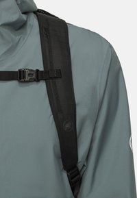 Grå texturerad jacka med ett svart rem, som har en sidspänne och en logotyp. Synliga sömmar och meshdetalj på axelområdet.