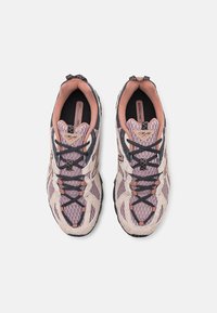 Zapatillas de trail running con parte superior de malla en rosa claro y beige, detalles en negro, suela texturizada y cordones contrastantes para un ajuste seguro.