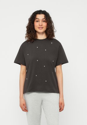 Γυναίκα με σγουρά μαλλιά, φοράει μια άνετη μαύρη μπλούζα με μικρά λευκά σχέδια σε σχήμα καρδιάς και ανοιχτογκρι παντελόνι, στέκεται μπροστά σε μονόχρωμο φόντο.