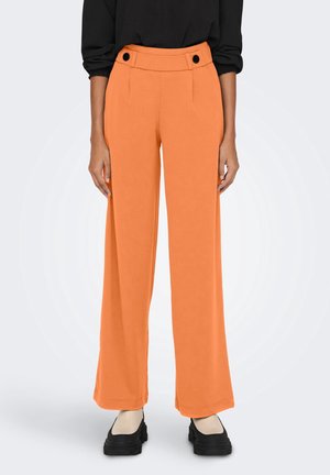 Kobieta nosząca pomarańczowe spodnie z wysokim stanem i szerokimi nogawkami z czarnymi guzikami, dobrane do czarnych butów na platformie i czarnego topu.