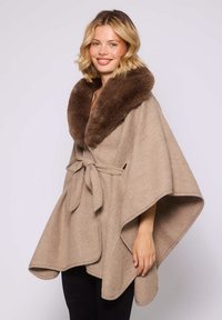 Femme souriante portant une cape en laine beige avec une ceinture nouée et un grand col en fourrure marron foncé, posant devant un fond clair uniforme.