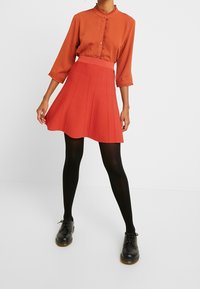 Blouse couleur rouille avec col à volants et détails à boutons, associée à une jupe trapèze rouge et des collants noirs, complétée par des chaussures noires.