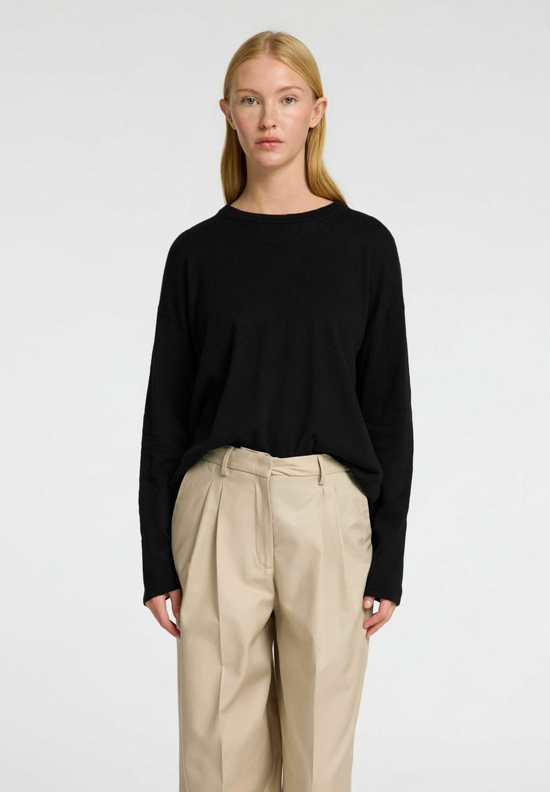 Černý svetr s dlouhým rukávem a kulatým výstřihem, doplněný o světle béžové kalhoty s vysokým pasem, které mají sklady a upravený střih.