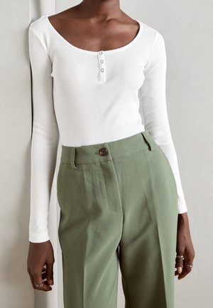 Femme portant un haut côtelé blanc à manches longues avec des boutons et un pantalon vert taille haute sur un fond neutre.
