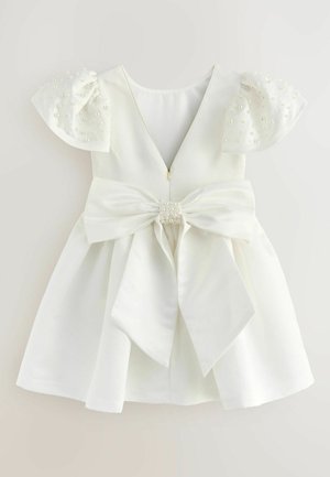 Biała sukienka z krótkimi, bufkowymi rękawami ozdobionymi małymi perłami. Posiada głęboki dekolt na plecach i dużą kokardę z przodu, mocowaną perłowym akcentem.