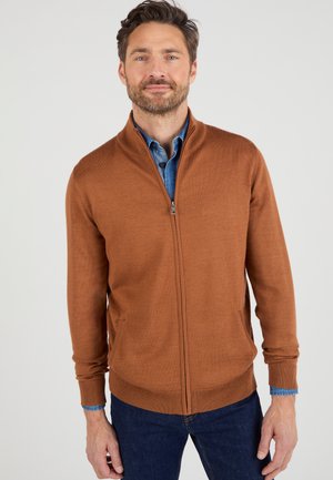 Bruine ritstrui van zacht weefsel, met een hoge kraag en zijzakken. Gedragen over een blauwe spijkerblouse.