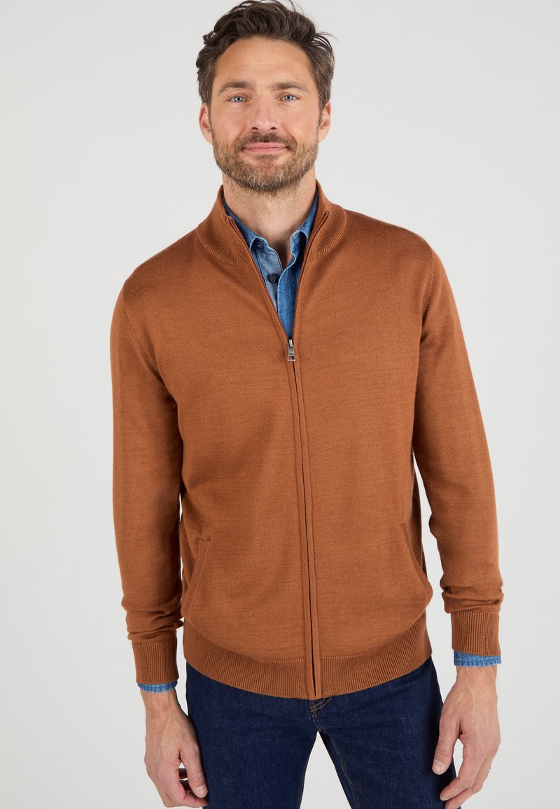 Suéter marrón con cremallera hecho de un suave tejido de punto, con un cuello alto y bolsillos laterales. Llevado sobre una camisa de mezclilla azul.