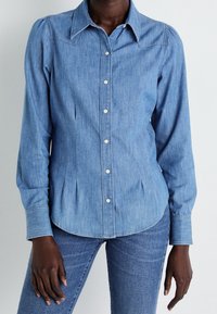 Ljusblå denimskjorta med spetsig krage, långa ärmar och knapplåsning. Har detaljer med smalveck och rundad fåll.