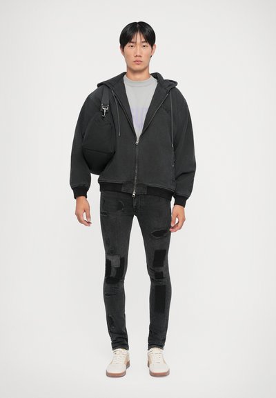 Čierna zip-up mikina s šedým grafickým tričkom pod ňou, opotrebované čierne džínsy s nášivkami a béžové tenisky.