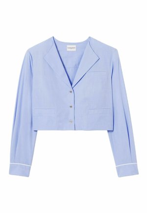 Mujer con blazer corto azul claro, falda plisada azul marino con doble cinturón, calcetines granate, zapatillas negras y lleva un bolso marrón.