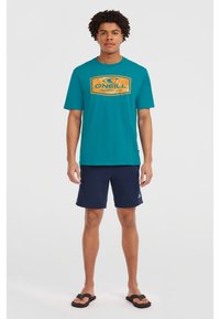 Camiseta de manga corta azul verdosa con un logo gráfico naranja, combinada con pantalones cortos azul marino. Llevada con chanclas negras. Diseño simple y casual.