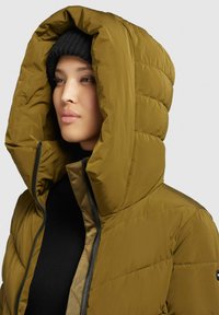 Giacca a vento in verde oliva con un grande cappuccio, chiusura con zip e texture trapuntata. Indossata sopra un maglione nero a maglia.