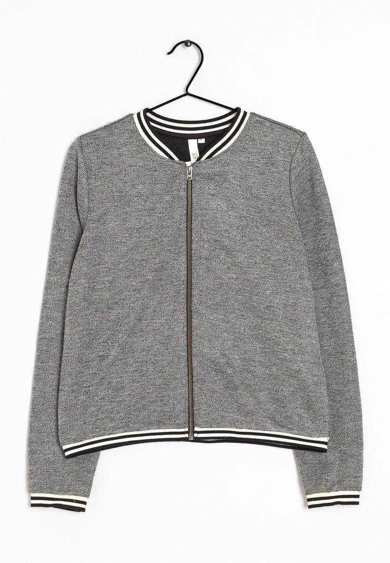 Veste grise zippée avec col, poignets et ourlet rayés noirs et blancs, suspendue sur un cintre noir contre un fond blanc.