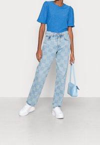 Osoba oblečená v modrém vzorovaném tričku s krátkým rukávem, světlomodrých kostkovaných džínách, bílých teniskách a držící malou světlomodrou kabelku.