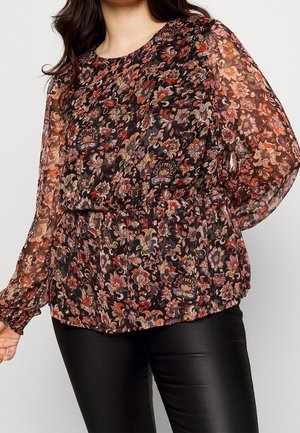 Vrouw met een doorschijnende blouse met lange mouwen en een meerkleurig bloemenpatroon, gecombineerd met zwarte leren broek.