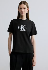 Svart bomulls-T-skjorte med en hvit "CK"-logo i fet skrift. Kortermet, rund hals og en ledig passform.