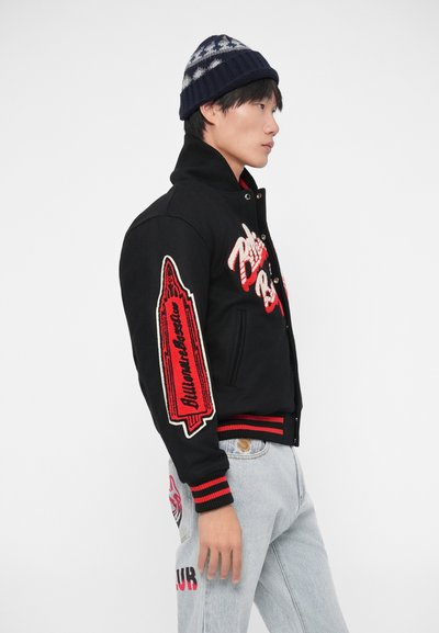 Casaco varsity preto com bordados em vermelho e creme, apresentando um design gráfico na manga. Combinado com jeans azul claro e um gorro estampado.