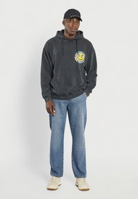 Felpa nera in cotone con tasca frontale e stampa grafica. Jeans in denim azzurro chiaro con vestibilità comoda, abbinati a scarpe da ginnastica beige e un cappellino nero.